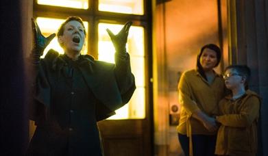 A dark image showing a woman dressed in a large coat telling ghost stories whilst a parent and older child listen.