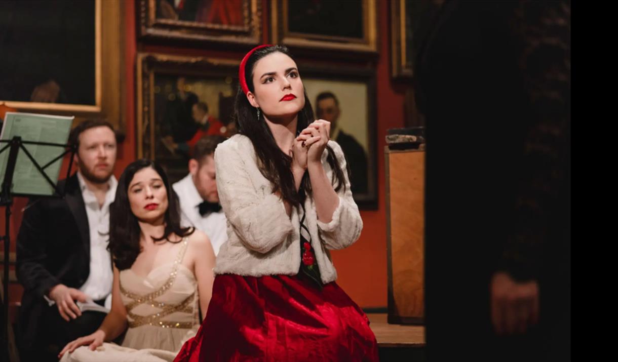 A woman in a red dress sitting on the floor holding her hands together. Behind her is a man in a suit and a woman in a gold dress.