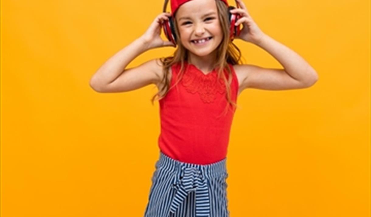 A young girl smiles and poses with her hands clasped to her headphones.
