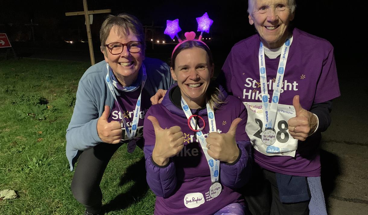 Three people smile and give thumbs up wearing Starlight Hike t-shirts, medals and flashing headwear, outdoors at Starlight Hike in Ferry Meadows.