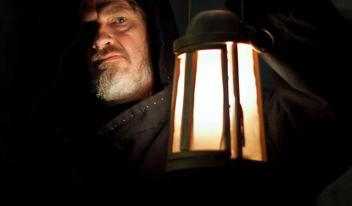 A man holding a lantern in the dark. Text reads: The Priestgate Vaults.