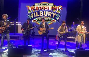 Roy Orbison & the Travelling Wilbury's performing on stage under blue lighting.