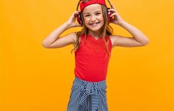 A young girl smiles and poses with her hands clasped to her headphones.