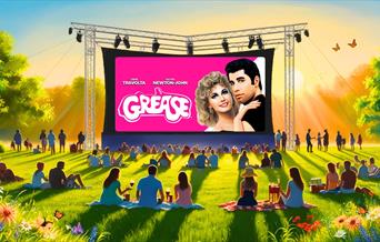 The film 'Grease' on a big screen outside with people on picnic blankets watching it.