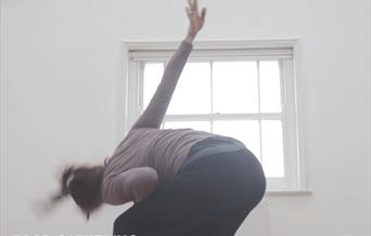 A dancer moves in front of a cream coloured wall and window