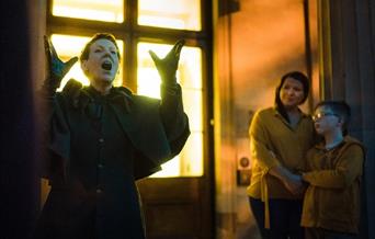 A dark image showing a woman dressed in a large coat telling ghost stories whilst a parent and older child listen.
