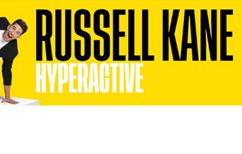 Russel Kane holding himself in the air with one hand against a yellow background.
