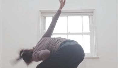 A dancer moves in front of a cream coloured wall and window