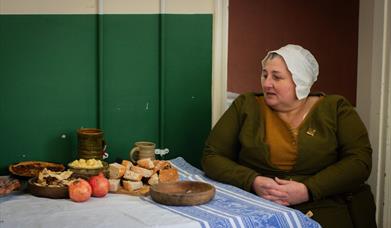 A woman in Tudor clothing, sitting at a table with a variety of Tudor period foods.