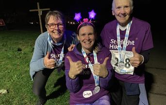 Three people smile and give thumbs up wearing Starlight Hike t-shirts, medals and flashing headwear, outdoors at Starlight Hike in Ferry Meadows.