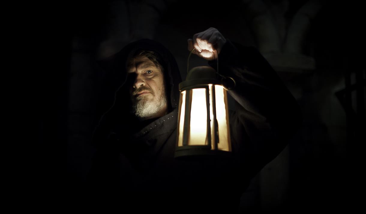 Cloaked figure holding a lantern in a darkened room
