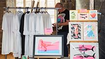 Display of white T-shirts on a rail beside colourful graphic art prints at an indoor market stall.