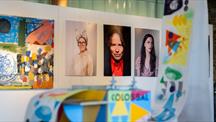 A colourful contemporary art exhibition: abstract paintings and three portrait photographs hang on a white gallery wall, partially framed by painted sculptural objects in the foreground.
