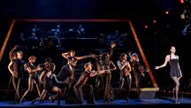 Dancers in black perform on stage with a live band behind them, while a solo woman in a sparkly dress stands to the side.
