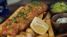A plate of golden battered fish and thick-cut chips, garnished with chopped parsley and served with a lemon wedge, mushy peas, and tartar sauce.

