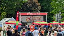 Crowds enjoy an outdoor “I Love Flavour Fest” live music stage in a sunny city square, with red and green parasols, food stalls and leafy trees creating a busy, festival atmosphere.