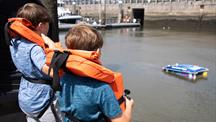 Two children in life jackets watch a small remote-controlled boat on the water.