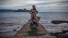 A woman in a trench coat stands on a concrete pier by the sea.