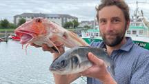 Man holding two fish up with a harbour in the background