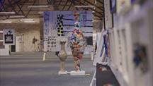 A large indoor art exhibition space featuring high ceilings with exposed wooden beams and bright overhead lighting. In the center foreground, two tall vases are displayed: one is plain and beige, while the other is colorful, covered in a patchwork of various patterns and designs. The background reveals several abstract artworks mounted on large panels, along with more art displayed on white walls and stands lining the right side of the image. The overall atmosphere is modern, creative, and spacious, with a focus on visual art installations and contemporary pieces.