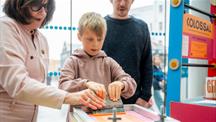 A woman is guiding a child as they both use a hands-on art or craft activity, with another adult standing behind them. The setting is indoors, brightly lit by natural light from large windows, and there are colorful signs and materials around, including a sign that reads "COLOSSAL." 