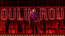 A man standing on a red stage with the word Moulin Rouge in lights behind