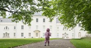 A little girl wearing a pink rainsuit runs towards Saltram House
