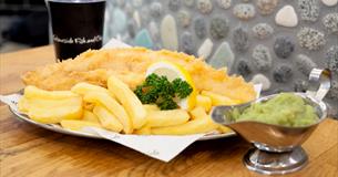 A plate of fish and chips garnished with lemon and parsley and a jug of mushy peas.