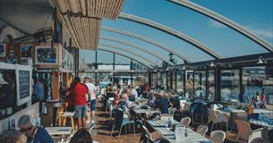 Inside the Boathouse Café with the roof retracted. Customers sitting at tables with a blue sky and views over Sutton Harbour.