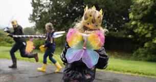 A child with rainbow fairy wings faces away from the camera in the foreground, with other children out of focus in the background.