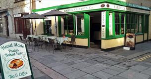 The green, white and yellow painted outside of the Barbican Pasty Co. with outside seating and tables and a sign.