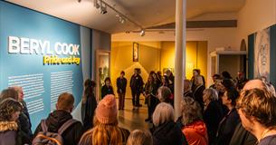 A large group of people listen to a talk in the 'Beryl Cook: Pride and Joy' exhibition at The Box Plymouth