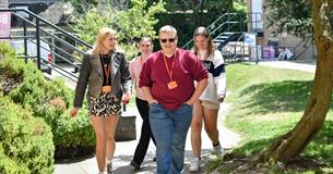 A group of four City College Plymouth students walk together outside on campus on a sunny day. They are smiling and wearing orange lanyards with ID ba