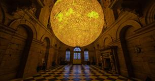 A glowing sun-like sphere hangs in the centre of an ornate hall with arched doorways, high ceilings, and a black-and-white checkered floor.
