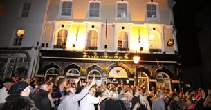 A crowd of people outside the brightly lit front of Kitty O'Hanlons at night-time.