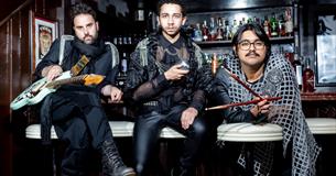Three men sit in front of a bar. Each hold a different thing: A guitar, a microphone and drumsticks. They wear black with silver embellishments and do