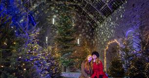 Two visitors sat on a bench in the Great Barn surrounded by Christmas trees covered in lights, a giant tree can be seen behind them.
