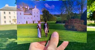 A hand holds a photograph showing two people in historical dress in front of a white estate, matching the real-life scene behind it.