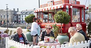 Red vintage Coffee and Donuts van with striped awning, outdoor seating area with Potted trees, white picket fence and customers sat at tables.