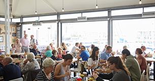 Busy inside of the restaurant showing tables full of diners in front of glass walls with views over Sutton Harbour.