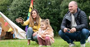 A family of three plays ring toss on grass near a tent with colourful flags; the mother and father kneel while the daughter watches.