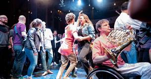 Image of 2 girls dancing together on the stage surrounded by other people, with people playing their instruments around them.