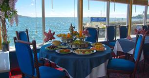 A table inside the restaurant set for a meal with dishes of food in front of large glass windows overlooking the water of Plymouth Sound.