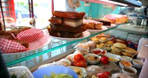 Large refrigerated display cabinet inside shop window displaying cakes, slices, pies and sandwich ingredients.