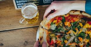 A large pizza divided into portions being pulled apart by a pair of hands. On the table is also a glass of beer and a menu.