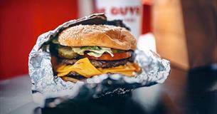 An opened silver foil package containing a large sesame seed topped bun containing a burger with salad and cheese.