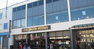 The glass frontage of the Union Rooms on a sunny day with a family and pushchair strolling by in the foreground.