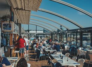 Inside the Boathouse Café with the roof retracted. Customers sitting at tables with a blue sky and views over Sutton Harbour.