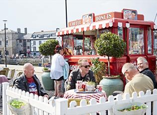 Red vintage Coffee and Donuts van with striped awning, outdoor seating area with Potted trees, white picket fence and customers sat at tables.