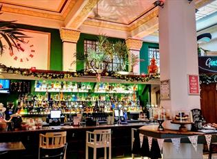 The bar area inside The Treasury, showing large stone pillars, large plants, a large wall clock and lots of bottles of drink displayed.